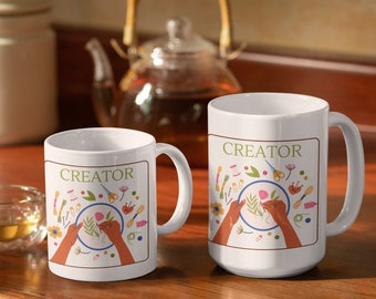 May include: White ceramic mugs with the word "CREATOR" in green, featuring a design of hands embroidering a floral pattern. The design includes scissors, thread, and other craft supplies. A teapot is in the background.