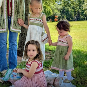 Op de afbeelding: Een familie in een parkomgeving, gekleed in handgemaakte haakkleding. Een vrouw draagt een lange groene cardigan met witte en roze randen, met een boek in haar hand. Drie jonge meisjes dragen gehaakte jurken in roze, wit en groen, met bijpassende handtassen. De jurken hebben een bloemenmotief.