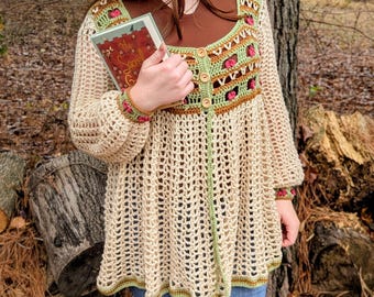 May include: A crochet beret shaped like a red apple with a green leaf and brown stem, paired with a beige crochet cardigan featuring a square neckline, floral embroidery, and three wooden buttons, worn over a brown shirt and blue jeans.