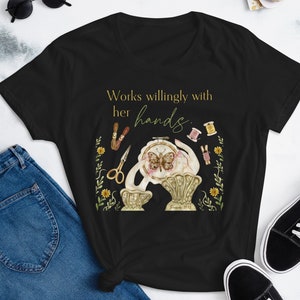 May include: Black t-shirt featuring a gold graphic of a hand holding a needle and thread with a butterfly on a hoop. The text "Works willingly with her hands." is printed in gold on the shirt.