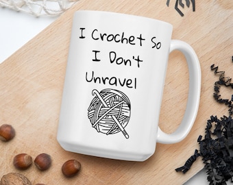 May include: White ceramic mug with black text and a drawing of a ball of yarn and crochet hook. The text reads "I crochet so I don't unravel."