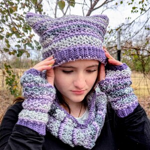 May include: A handmade crochet cat ear hat, fingerless gloves, and cowl in shades of purple, gray, and white. The hat has pointed ears and a ribbed brim. The gloves and cowl have a matching pattern. The set is displayed outdoors.