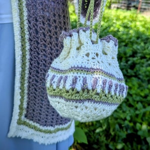 May include: A cream-colored, crocheted drawstring bag with green and purple accents. The bag is held by green cords and is next to a matching crocheted shawl with a purple and green design. The background is blurred green foliage.