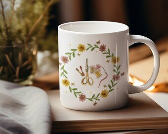 May include: White ceramic mug with a floral wreath design featuring a pair of scissors, thread spools, and needles. The wreath is made of yellow and pink flowers with green leaves.