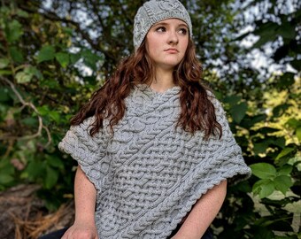 May include: A woman wearing a gray knitted poncho with a cable knit pattern and a gray knitted hat. She is sitting on a log in a wooded area.