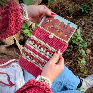 May include: A handmade crochet book sleeve in shades of red, green, and white, with a button closure. The sleeve is holding a book with the title "The". The crochet sleeve is held by a person wearing a red crochet top.