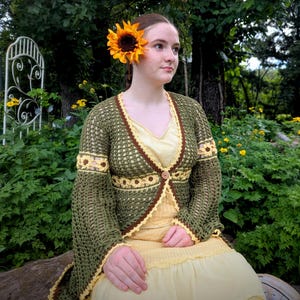 Pode incluir: Uma mulher usando um cardigã de crochê verde feito à mão com detalhes marrons e amarelos, combinado com um vestido amarelo. Um grande girassol é colocado em seu cabelo. A cena é ao ar livre com vegetação ao fundo.