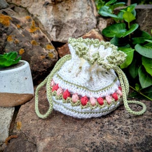 May include: A white crochet drawstring bag with a green trim and strap. The bag features a band of pink and peach-coloured floral embroidery. A small succulent plant in a white and brown pot sits nearby on a stone surface.