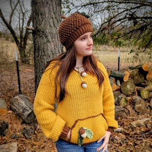 May include: A mustard yellow crochet hoodie with a brown trim and a front pocket adorned with an acorn. The outfit includes a matching brown acorn hat with a green leaf accent. The hat and hoodie are handmade.