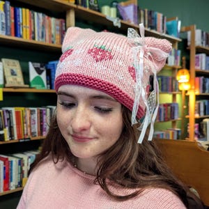 May include: A light pink knit beanie with cat ears, featuring a strawberry pattern, a dark pink ribbed brim, and a white sheer ribbon bow attached to one side. The beanie is paired with a matching light pink knit sweater with a strawberry design.