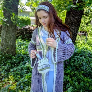 May include: A handmade crochet ensemble in shades of purple, white, and green. The set includes a long cardigan, headband, and a small drawstring bag. The cardigan features bell sleeves and decorative trim. The ensemble is perfect for a bohemian or cottagecore aesthetic.