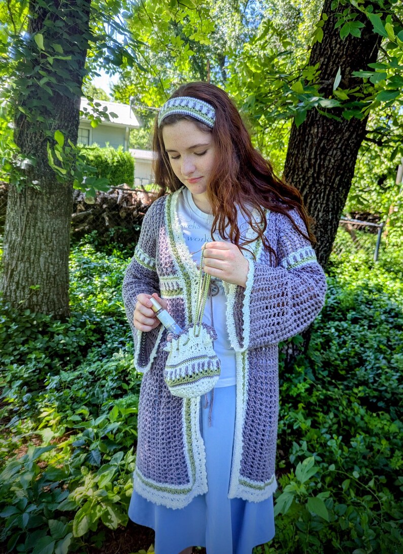 May include: A handmade crochet ensemble in shades of purple, white, and green. The set includes a long cardigan, headband, and a small drawstring bag. The cardigan features bell sleeves and decorative trim. The ensemble is perfect for a bohemian or cottagecore aesthetic.