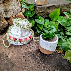 May include: A white crochet drawstring bag with green trim and a pink and red floral pattern. A small white and brown ceramic pot holds a green succulent plant. The items are on a stone surface.