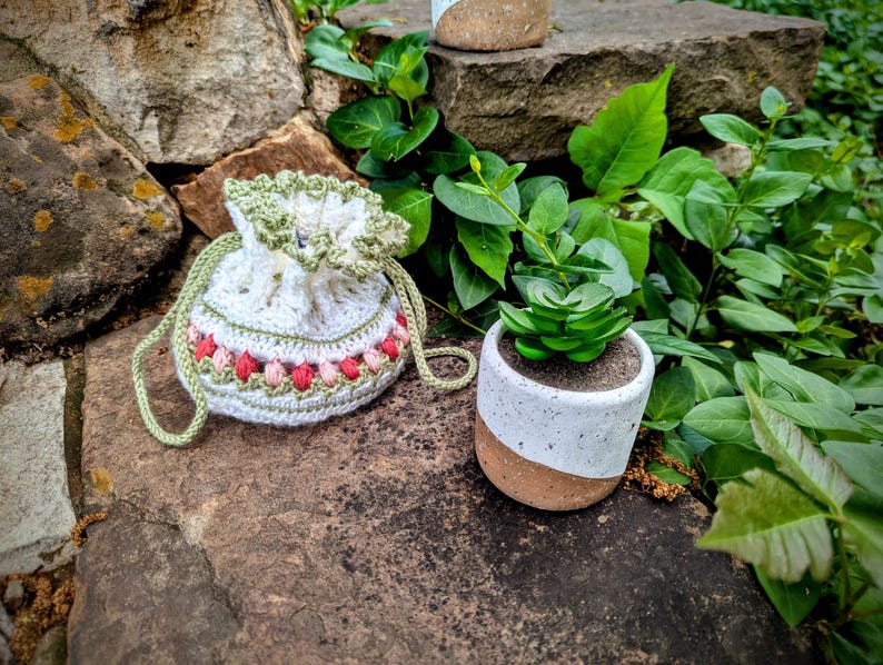May include: A white crochet drawstring bag with green trim and a pink and red floral pattern. A small white and brown ceramic pot holds a green succulent plant. The items are on a stone surface.