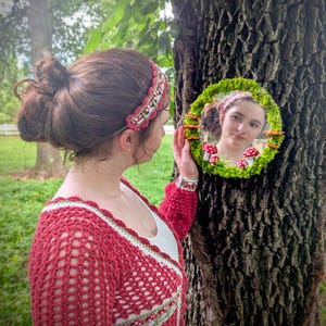 May include: A woman in a red crochet sweater and headband, holding a round mirror decorated with moss and mushrooms. The mirror reflects the woman's face and the forest setting. The headband and sweater have a green and white trim.