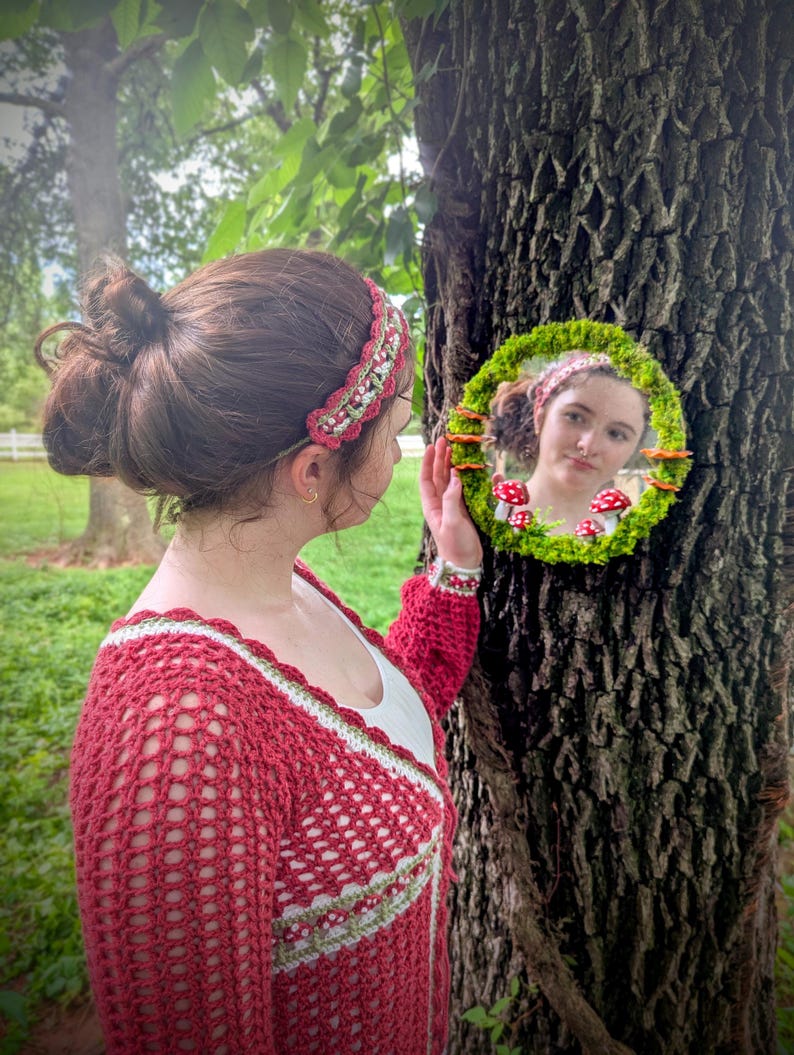 May include: A woman in a red crochet sweater and headband, holding a round mirror decorated with moss and mushrooms. The mirror reflects the woman's face and the forest setting. The headband and sweater have a green and white trim.