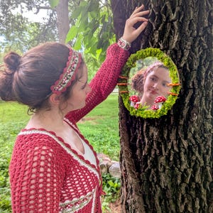 May include: An oval mirror, framed with moss and small mushrooms, is attached to a tree. The mirror reflects a person. The person wears a red crochet cardigan and a matching headband over a white top.