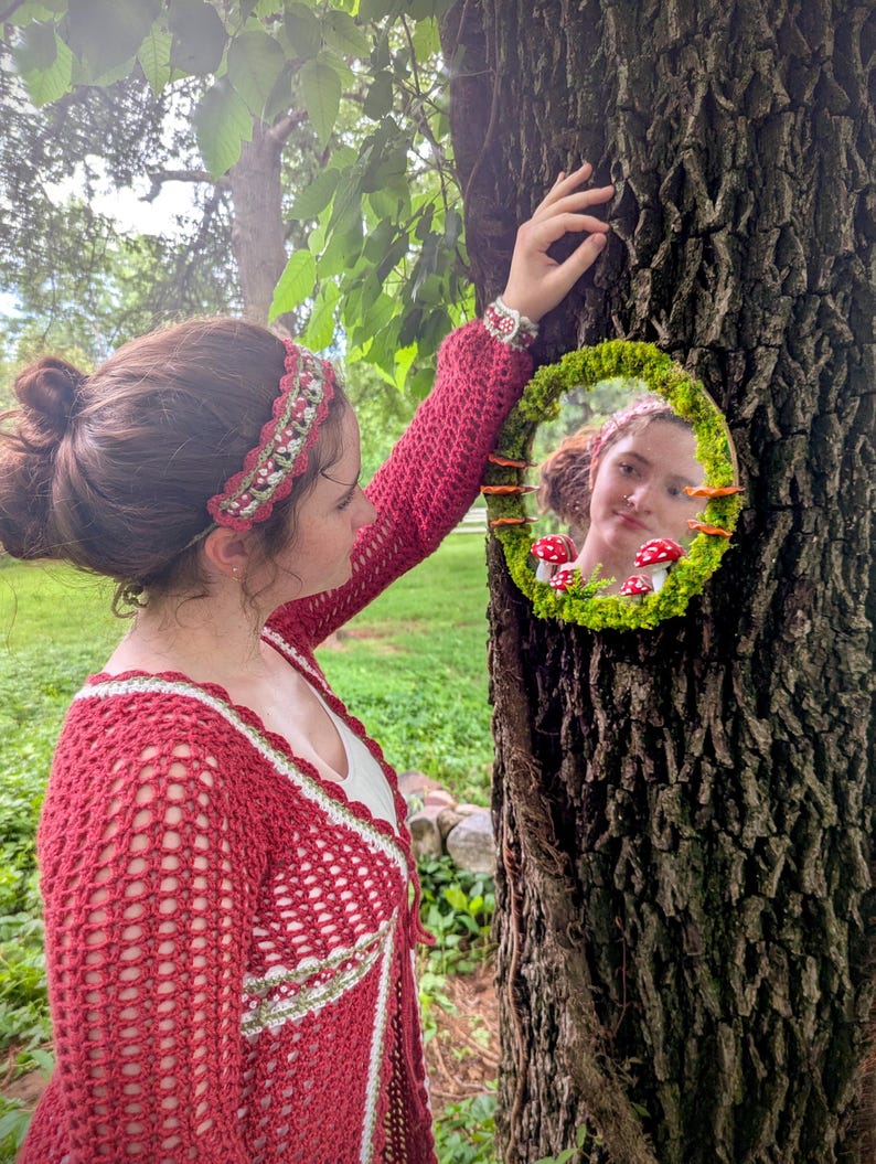 May include: An oval mirror, framed with moss and small mushrooms, is attached to a tree. The mirror reflects a person. The person wears a red crochet cardigan and a matching headband over a white top.