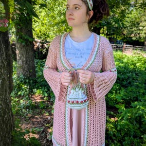 Op de afbeelding: Een lichtroze, opengebreid gehaakt vest met witte en groene randen. Het vest wordt gedragen over een wit t-shirt en een roze jurk. Een bijpassende haarband en een kleine handtas met bloemendetails maken de outfit compleet.