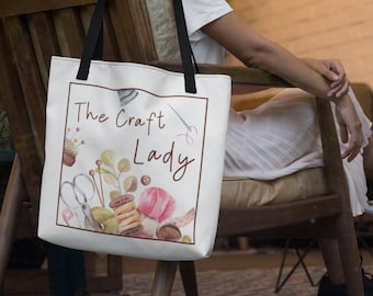 May include: White tote bag with black handles featuring the text "The Craft Lady" in brown script. The design includes watercolor illustrations of sewing tools, yarn, and other craft supplies within a brown border.