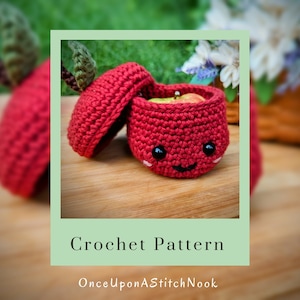 May include: A crocheted apple-shaped box with a lid, featuring a smiling face and black eyes. The box is red and has a green leaf on top. The box is open and the inside is visible. The box is sitting on a wooden surface. Crochet Pattern OnceUponAStitchNook