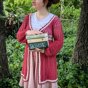 May include: A red crochet cardigan with white trim is worn over a white t-shirt and pink dress. A matching headband and a stack of books are also visible. The background is a lush green forest.