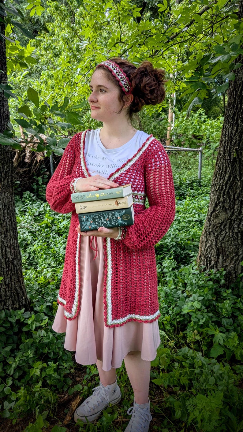 May include: A red crochet cardigan with white trim is worn over a white t-shirt and pink dress. A matching headband and a stack of books are also visible. The background is a lush green forest.