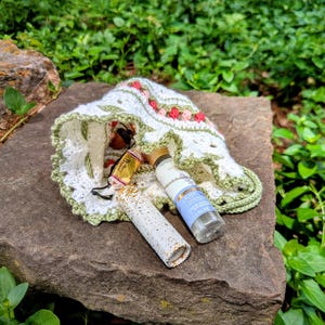 May include: A white crochet hat with pink and red floral accents and a green trim, resting on a stone surface. Beside the hat are two small bottles and a tube with gold accents. The background is filled with green foliage.