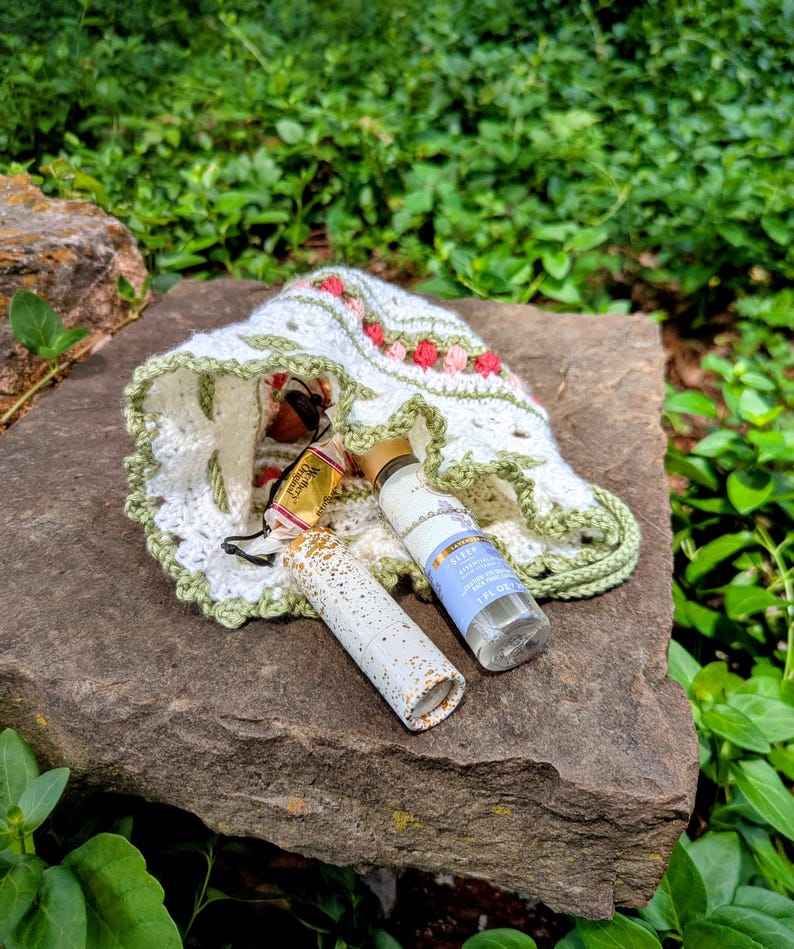 May include: A white crochet hat with pink and red floral accents and a green trim, resting on a stone surface. Beside the hat are two small bottles and a tube with gold accents. The background is filled with green foliage.
