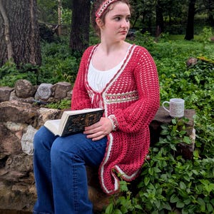 May include: A red crochet cardigan with white trim is worn over a white top and blue jeans. A matching headband and bracelet are also visible. The person is holding a book with gold lettering. A mug sits on a stone surface.