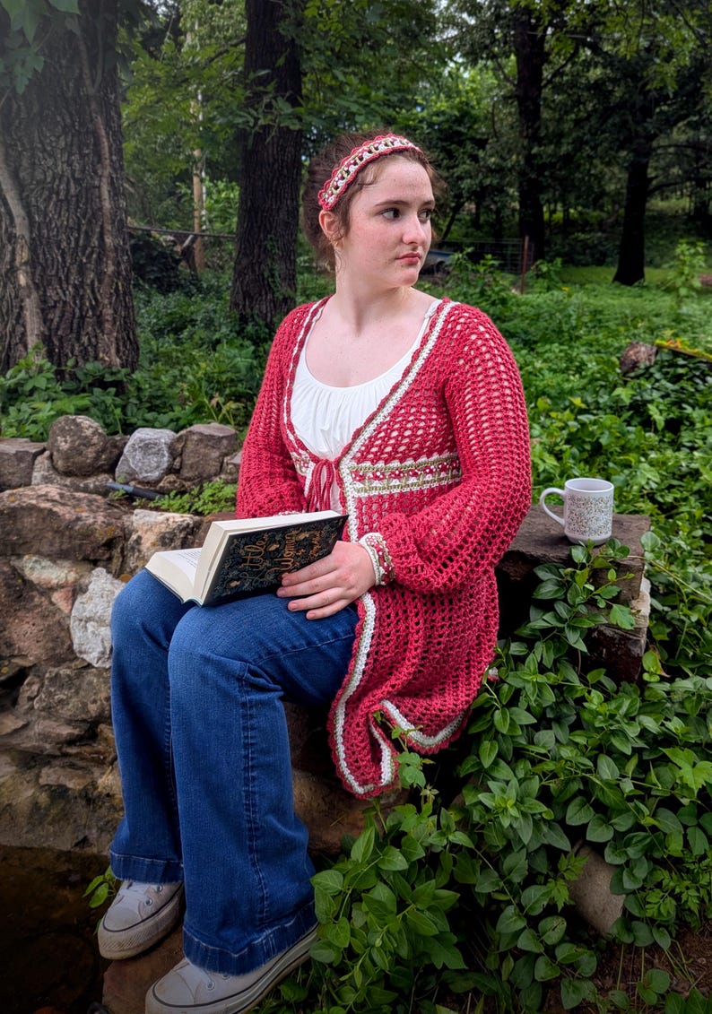 May include: A red crochet cardigan with white trim is worn over a white top and blue jeans. A matching headband and bracelet are also visible. The person is holding a book with gold lettering. A mug sits on a stone surface.