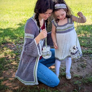 May include: A mother and child wearing matching crochet outfits. The mother wears a long, open-front cardigan in gray with white and green trim, paired with jeans. The child wears a white dress with gray and green accents, a matching headband, and a small purse.