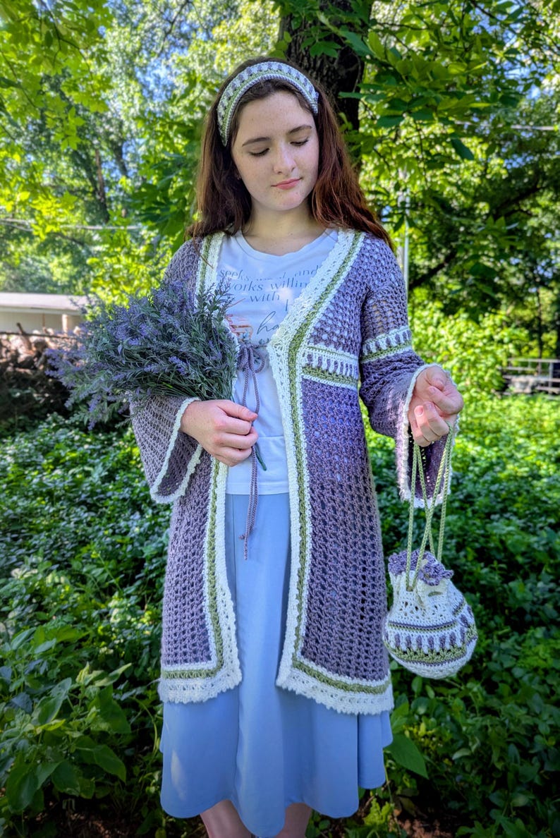 May include: A handmade crochet ensemble in shades of purple, white, and green. The outfit includes a long cardigan, a small drawstring bag, and a matching headband. The cardigan features a delicate open weave and trim. The ensemble is perfect for a bohemian or cottagecore aesthetic.