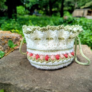 May include: A white crochet drawstring bag with green and red floral accents. The bag features a scalloped green trim and green drawstring handles. The bag is sitting on a gray stone surface with a green background.