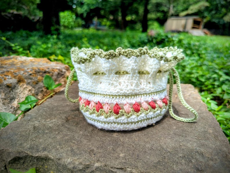 May include: A white crochet drawstring bag with green and red floral accents. The bag features a scalloped green trim and green drawstring handles. The bag is sitting on a gray stone surface with a green background.