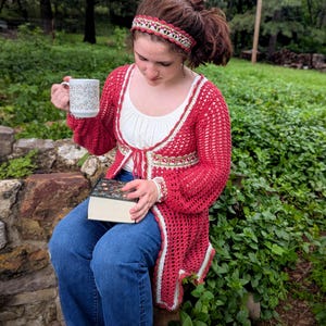 May include: A red crochet cardigan with white trim, and a matching headband. The cardigan is worn over a white top and blue jeans. A floral-patterned mug and a book are also visible.