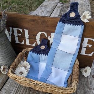May include: Two blue and white checkered kitchen towels with navy blue crocheted tops and wooden buttons. The towels are hanging on a wooden sign that says "WELCOME".