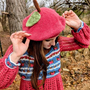 May include: A handmade, red crochet beret with a brown stem and green leaf accent. The beret is styled with a matching red crochet dress with blue and white accents. The dress has long sleeves and a unique openwork design.