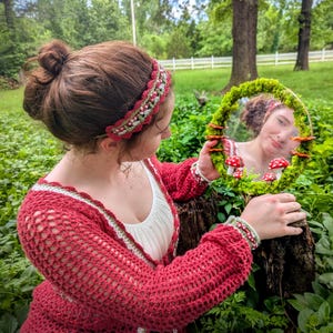 May include: A handmade red crochet cardigan with white and green trim, and a matching headband. The person is holding a round mirror decorated with moss and mushroom accents. The mirror reflects the person's face.