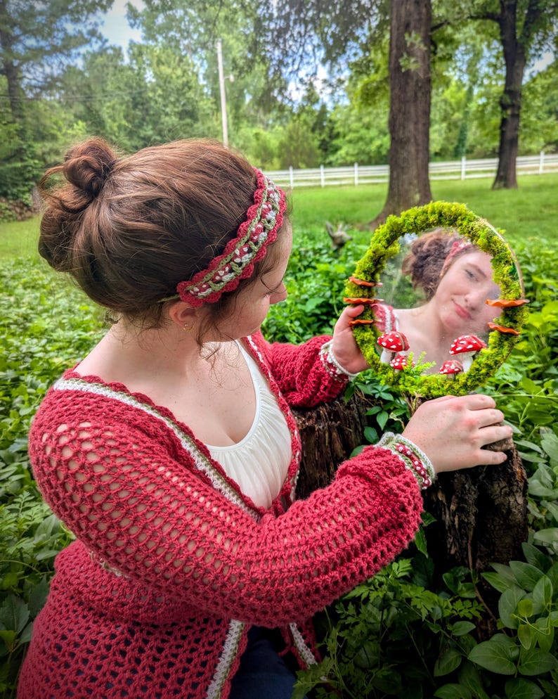 May include: A handmade red crochet cardigan with white and green trim, and a matching headband. The person is holding a round mirror decorated with moss and mushroom accents. The mirror reflects the person's face.