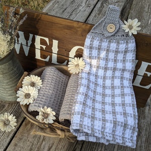 May include: A white and gray checkered kitchen towel with a crocheted top and a wooden button. The towel is hanging on a wooden sign that says "WELCOME".