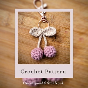 May include: A crochet cherry keychain with a pink and beige color scheme. The keychain features a small bow and a gold key ring. The text "Crochet Pattern" and "OnceUponAStitchNook" is displayed below the image.