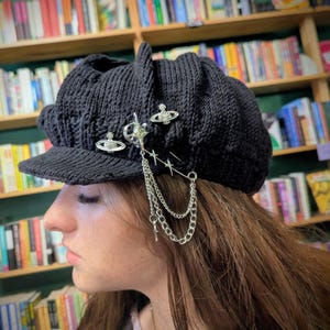 May include: A black knitted newsboy cap featuring a stiff brim and a textured ribbed pattern. The hat is adorned with three silver-toned metal pins, including two orb-shaped emblems and a central star-shaped charm connected by delicate silver chains.