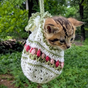 May include: A small tabby kitten peeks out of a white crochet bag with a green trim and strap. The bag is decorated with red and pink floral patterns. The background is a blurred view of green foliage.