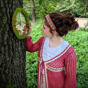 May include: A red crochet cardigan with white and green trim, and a matching headband. The image also features a round mirror decorated with moss and miniature mushrooms, creating a whimsical, nature-inspired aesthetic.