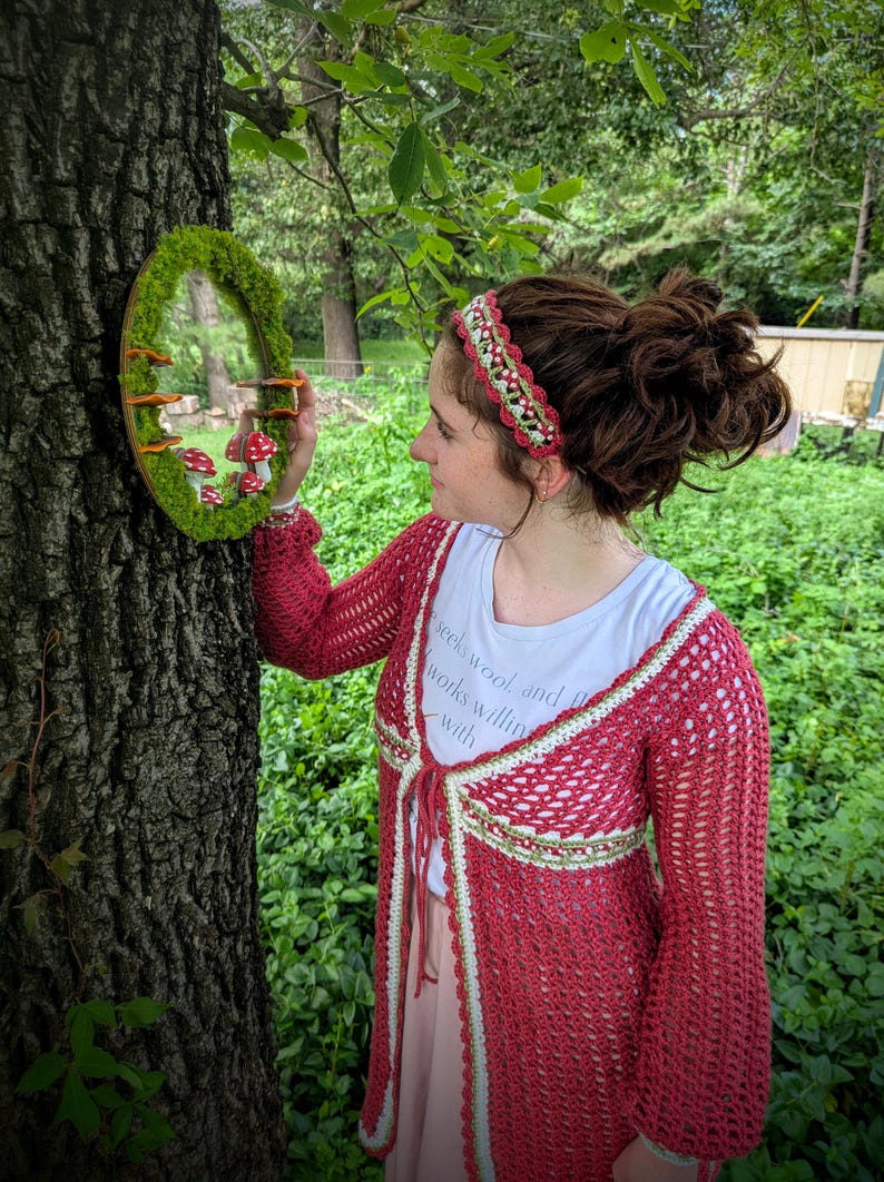May include: A red crochet cardigan with white and green trim, and a matching headband. The image also features a round mirror decorated with moss and miniature mushrooms, creating a whimsical, nature-inspired aesthetic.