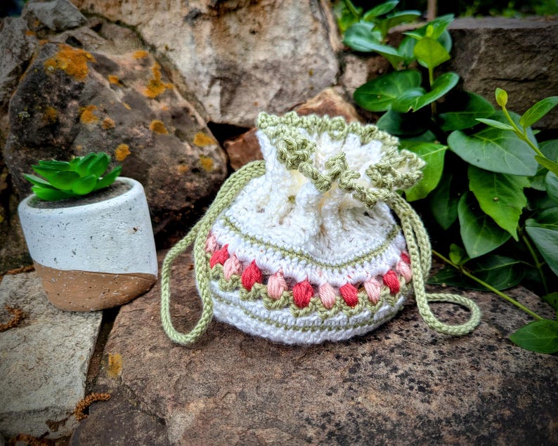 May include: A white crochet drawstring bag with a green trim and strap. The bag features a band of pink and peach-colored floral embroidery. A small succulent plant in a white and brown pot sits nearby on a stone surface.