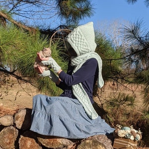May include: A woman wearing a light green hooded scarf and matching fingerless gloves sits on a pile of logs in a wooded area. She is holding a small kitten in her hands.
