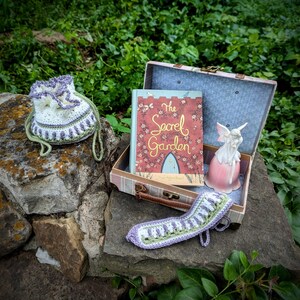 May include: A crochet drawstring bag, a book titled "The Secret Garden", and a decorative fairy figurine are displayed on a stone surface. The book is in a vintage suitcase. A crochet item is also present.