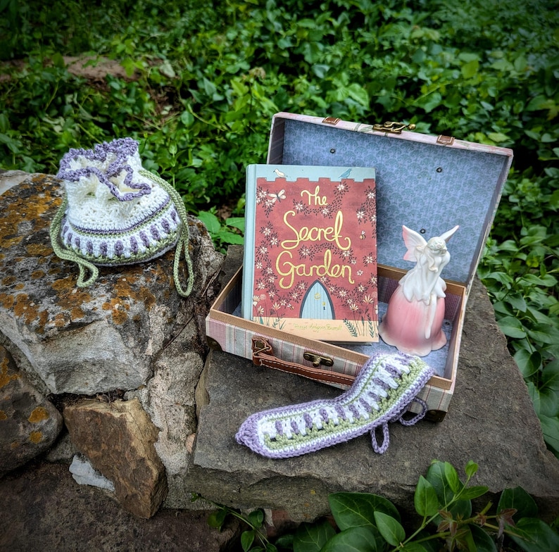May include: A crochet drawstring bag, a book titled "The Secret Garden", and a decorative fairy figurine are displayed on a stone surface. The book is in a vintage suitcase. A crochet item is also present.